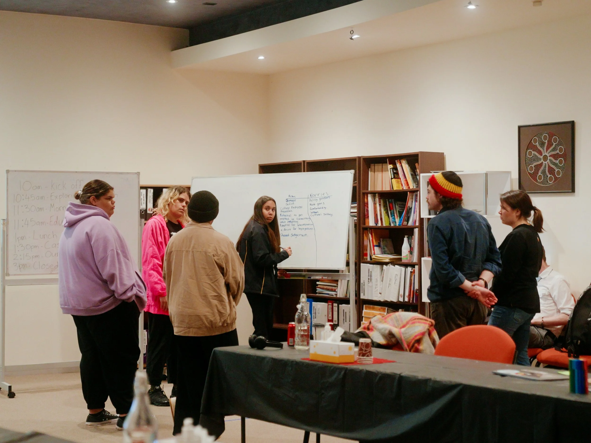 A group of people standing in a circle in a room, with a whiteboard and bookshelves in the background, engaging in a discussion or activity.