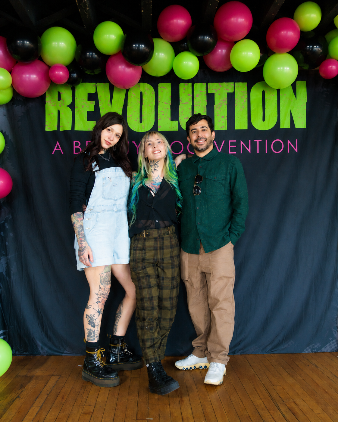 Three people standing in front of a black backdrop decorated with pink, green, and black balloons, and the word 'REVOLUTION' in large green letters, with pink text underneath.