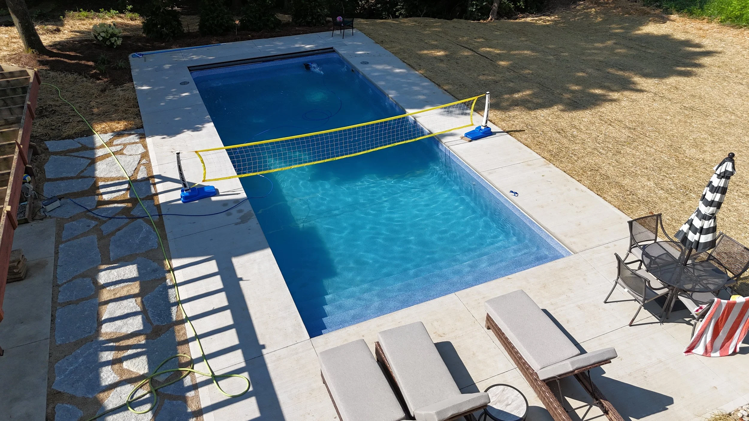 Swimming pool with a volleyball net, pool cleaning equipment, and outdoor furniture including lounge chairs, a table, and a striped umbrella.