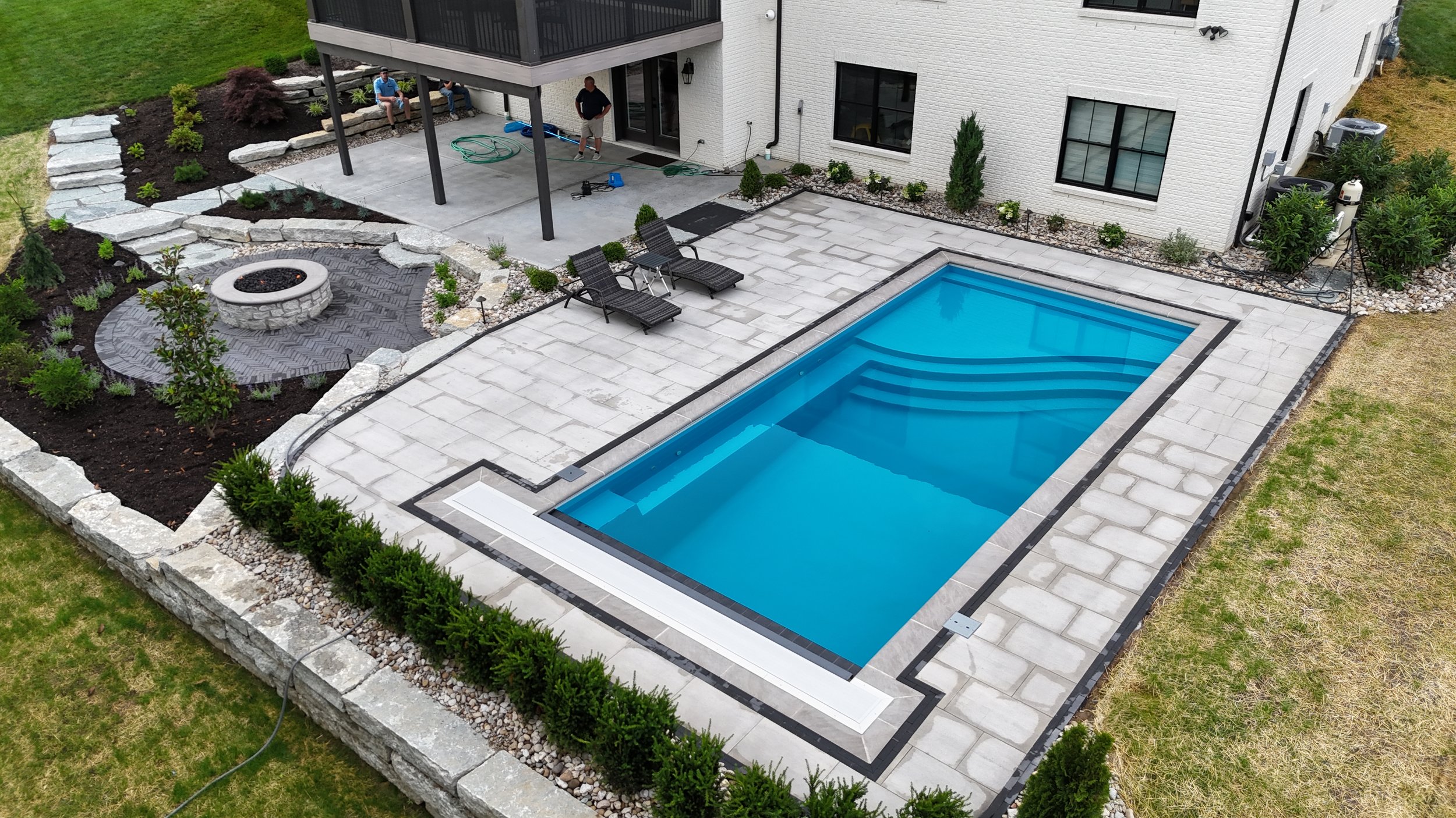 A backyard with a swimming pool, a sitting area with two lounge chairs, and a fire pit area surrounded by landscaping and a multi-story white house.
