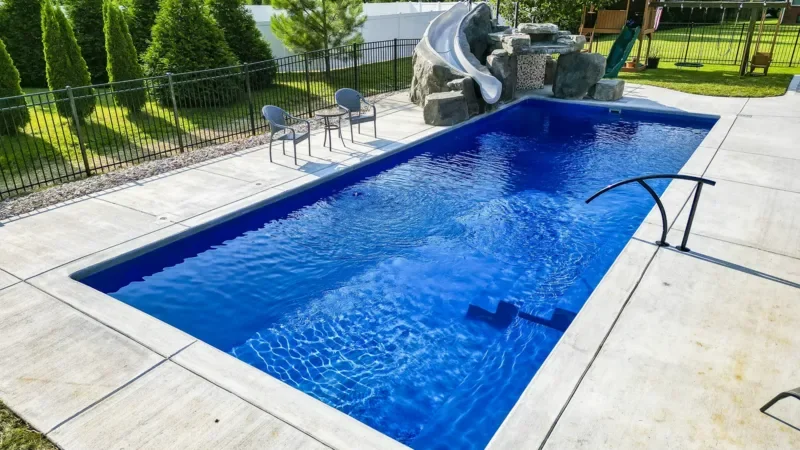 In-ground rectangular fibergladd swimming pool in mason, oh with a small attached water slide and rocks, surrounded by concrete patio and black metal fence, with two chairs nearby.