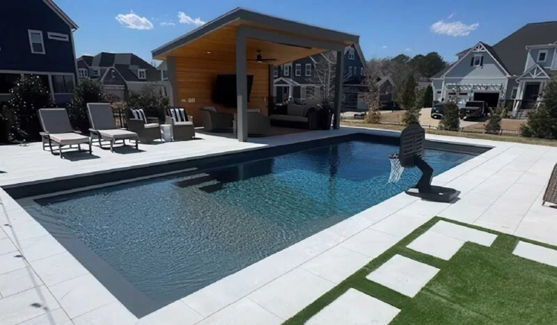 Residential backyard with a fiberglass swimming pool in west harrison, IN, lounge chairs, a covered outdoor living area, and neighboring houses under a partly cloudy sky.