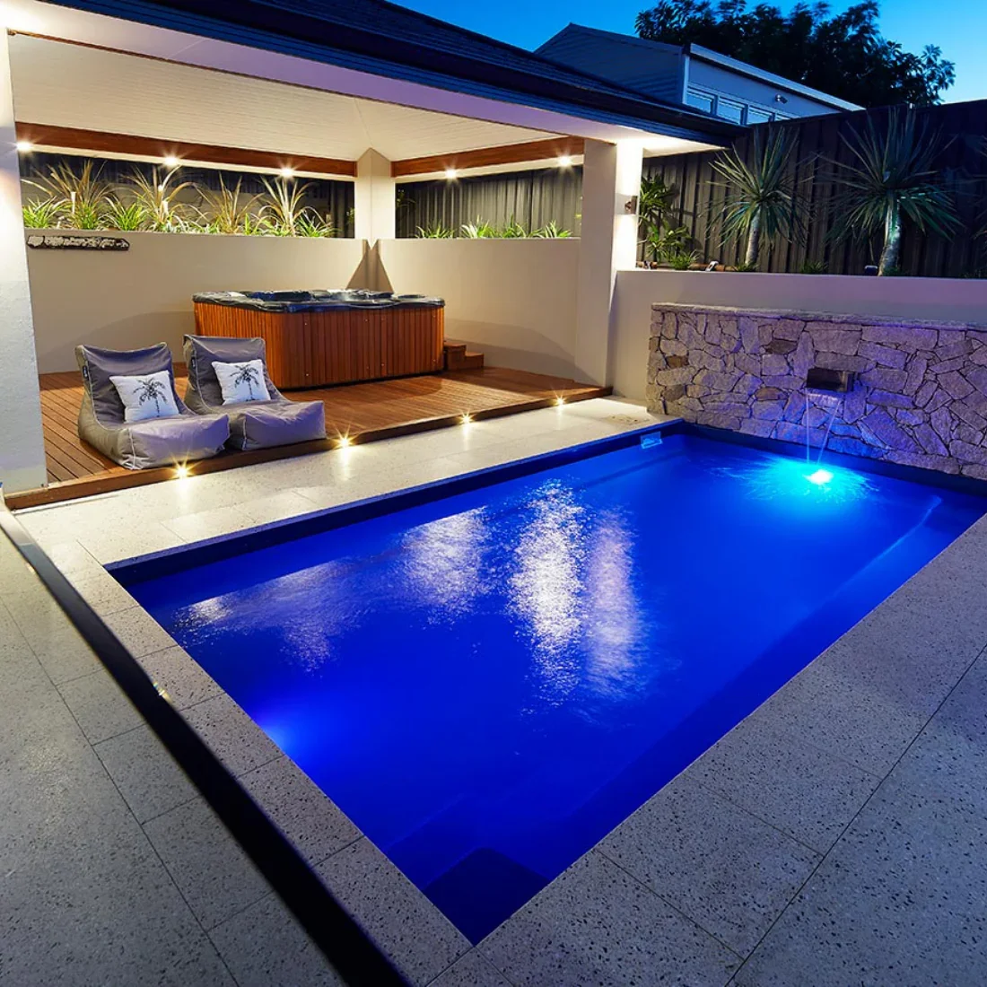 An outdoor backyard with a small illuminated swimming pool, a hot tub, outdoor seating with cushions, and tropical plants under a covered patio during evening. Located in Blue Ash, OH