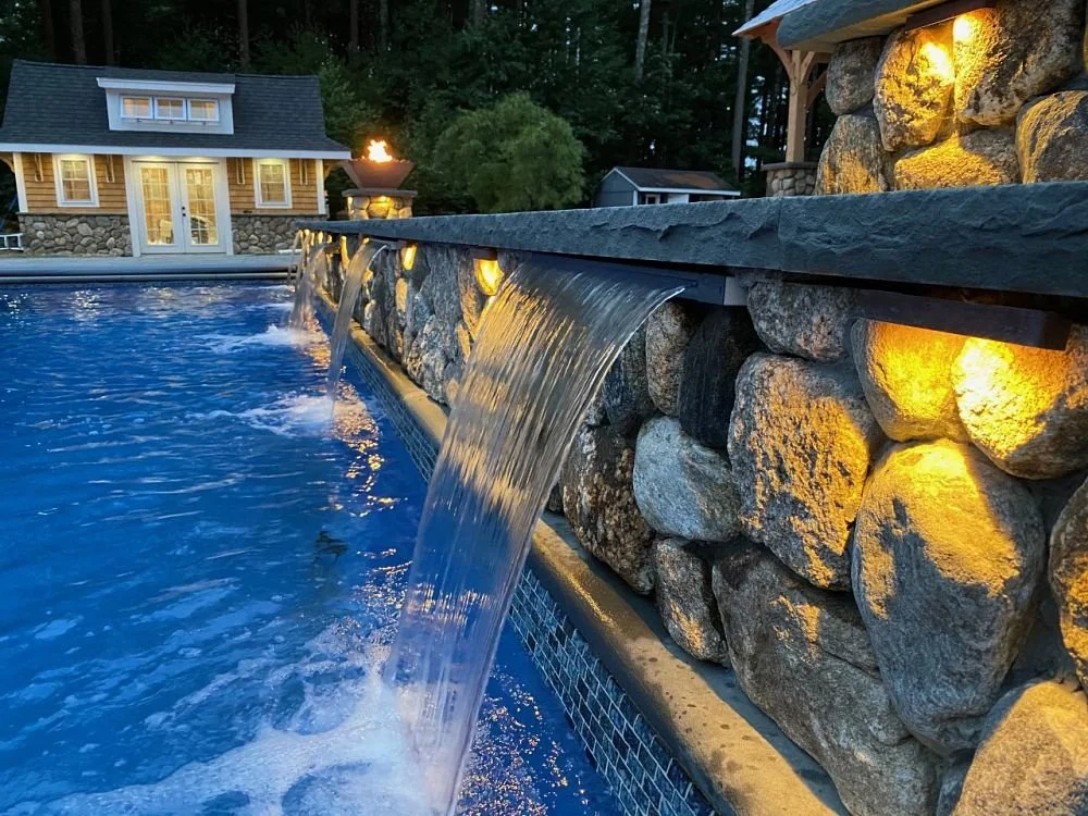 A stone waterfall feature with water cascading into a pool at dusk, illuminated by warm yellow lights, with a cozy house and trees in the background.