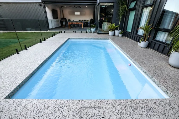 Rectangular outdoor fiberglass swimming pool with a speckled stone deck, surrounded by potted plants and adjacent to a modern patio with large sliding glass doors.