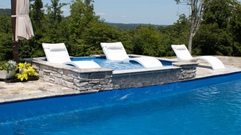 Poolside lounge chairs in a shallow water area above an fiberglass infinity pool, surrounded by trees.