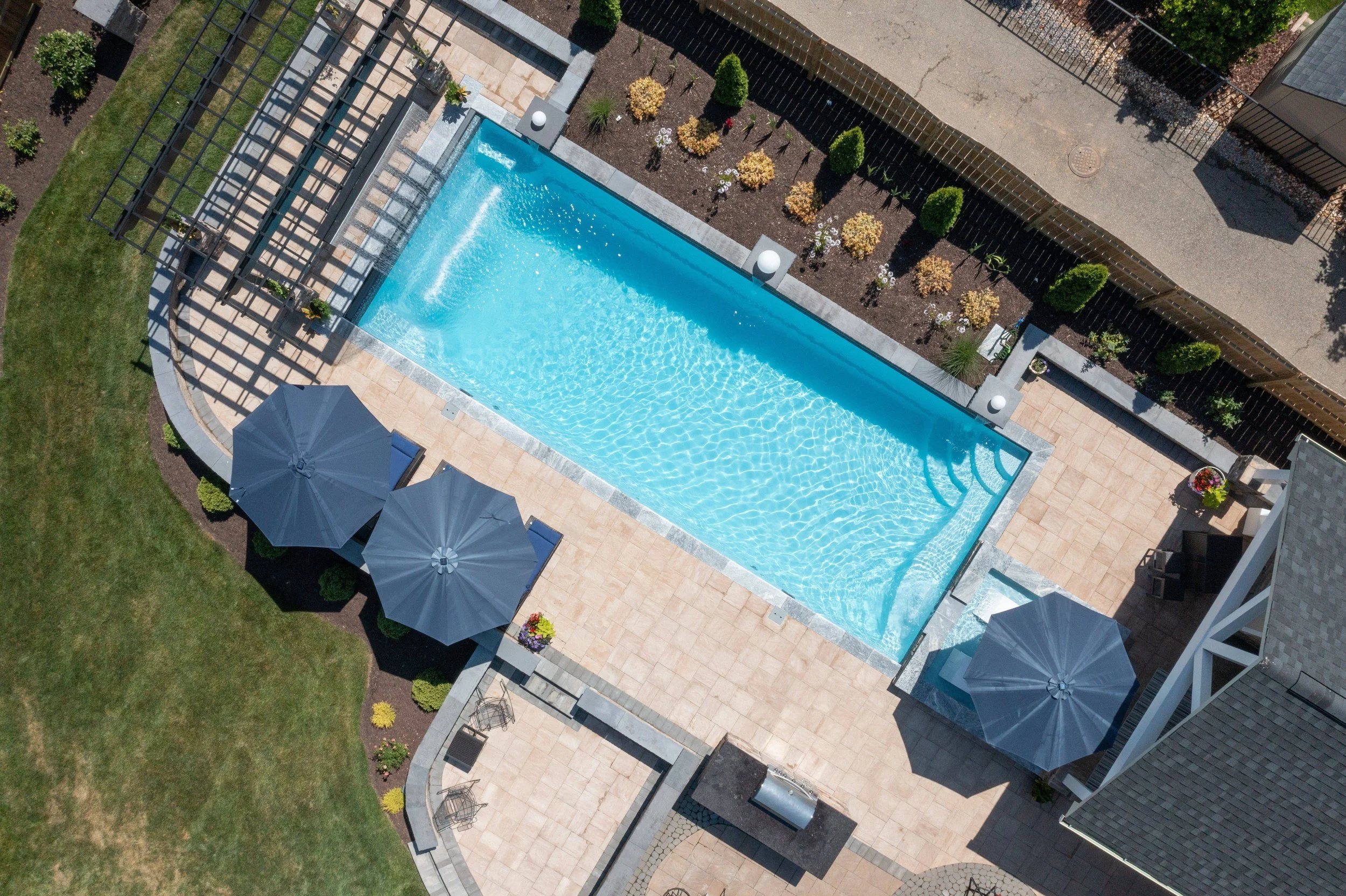 Aerial view of a rectangular swimming pool with clear blue water, surrounded by a paved deck with umbrellas, chairs, and landscaped garden with bushes and flowers, enclosed by a fence. In Petersburg, KY