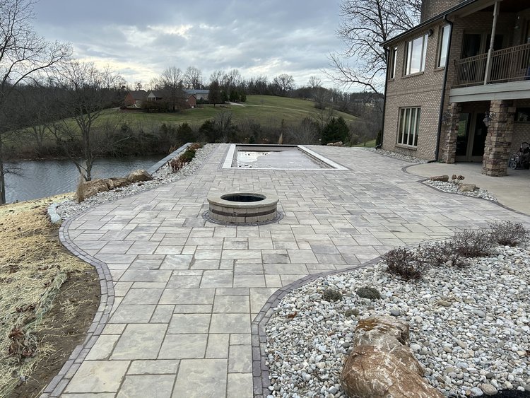 A newly constructed patio with light gray paving stones, a fire pit, and a rectangular pool near a house with a lawn and trees in the background.