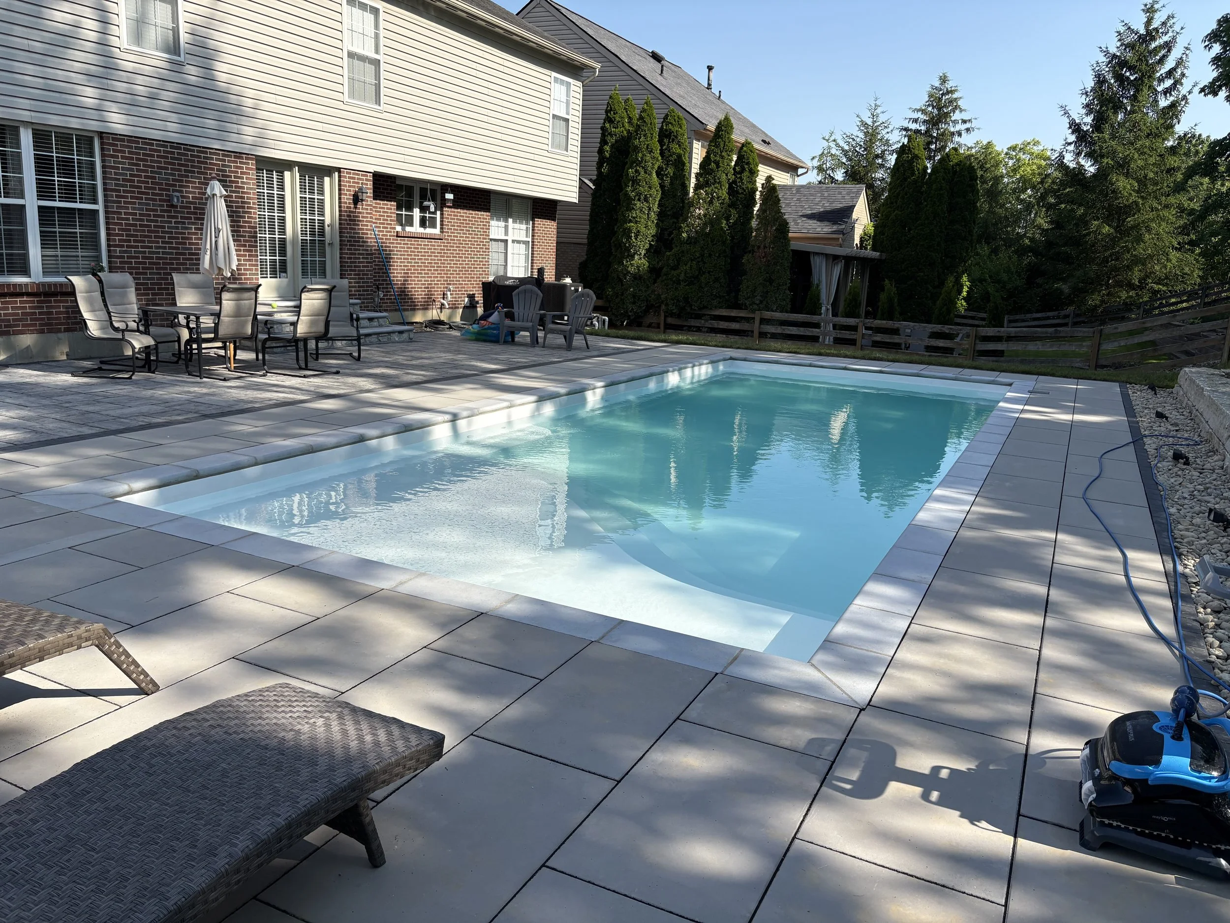Backyard with in-ground swimming pool, patio furniture, and a house with brick and siding exterior, surrounded by trees and a wooden fence.