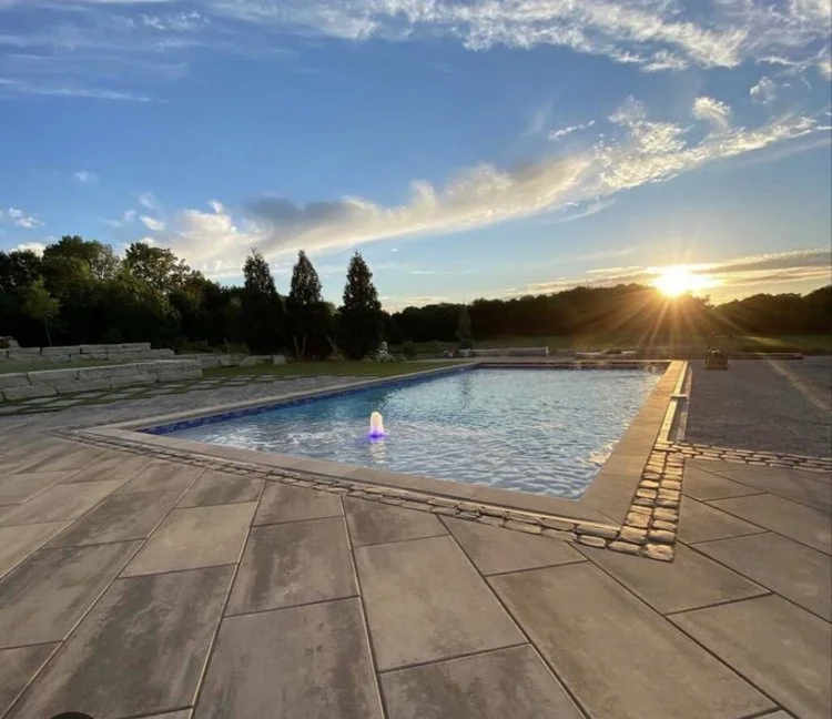 Outdoor swimming pool with a fountain, surrounded by stone pavement, with the sun setting in the background and trees in the distance.