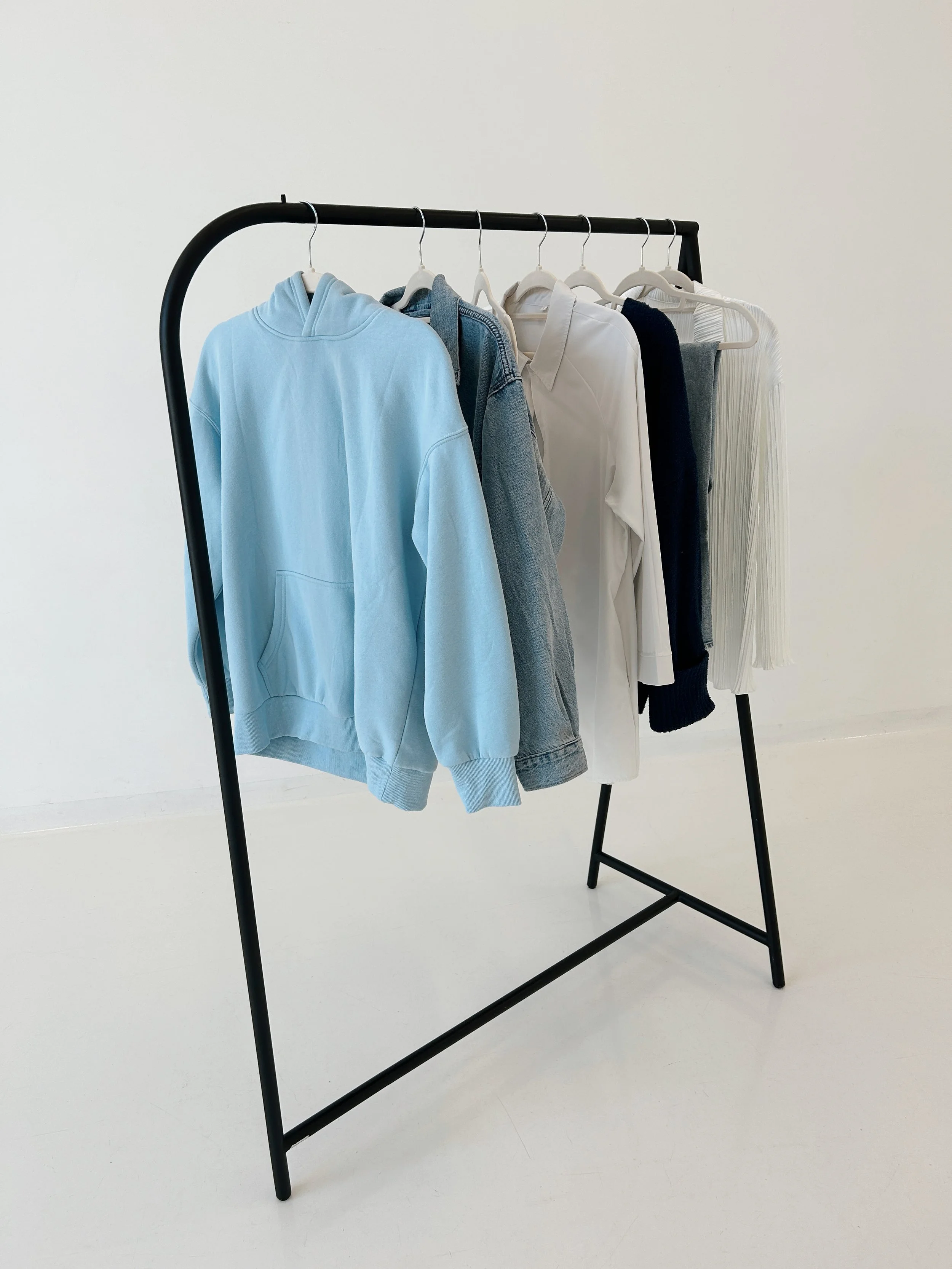 Clothing rack with various pieces of clothing including a light blue hoodie, denim jacket, white shirt, dark sweater, gray tank top, and a white pleated skirt on white hangers against a plain white background.