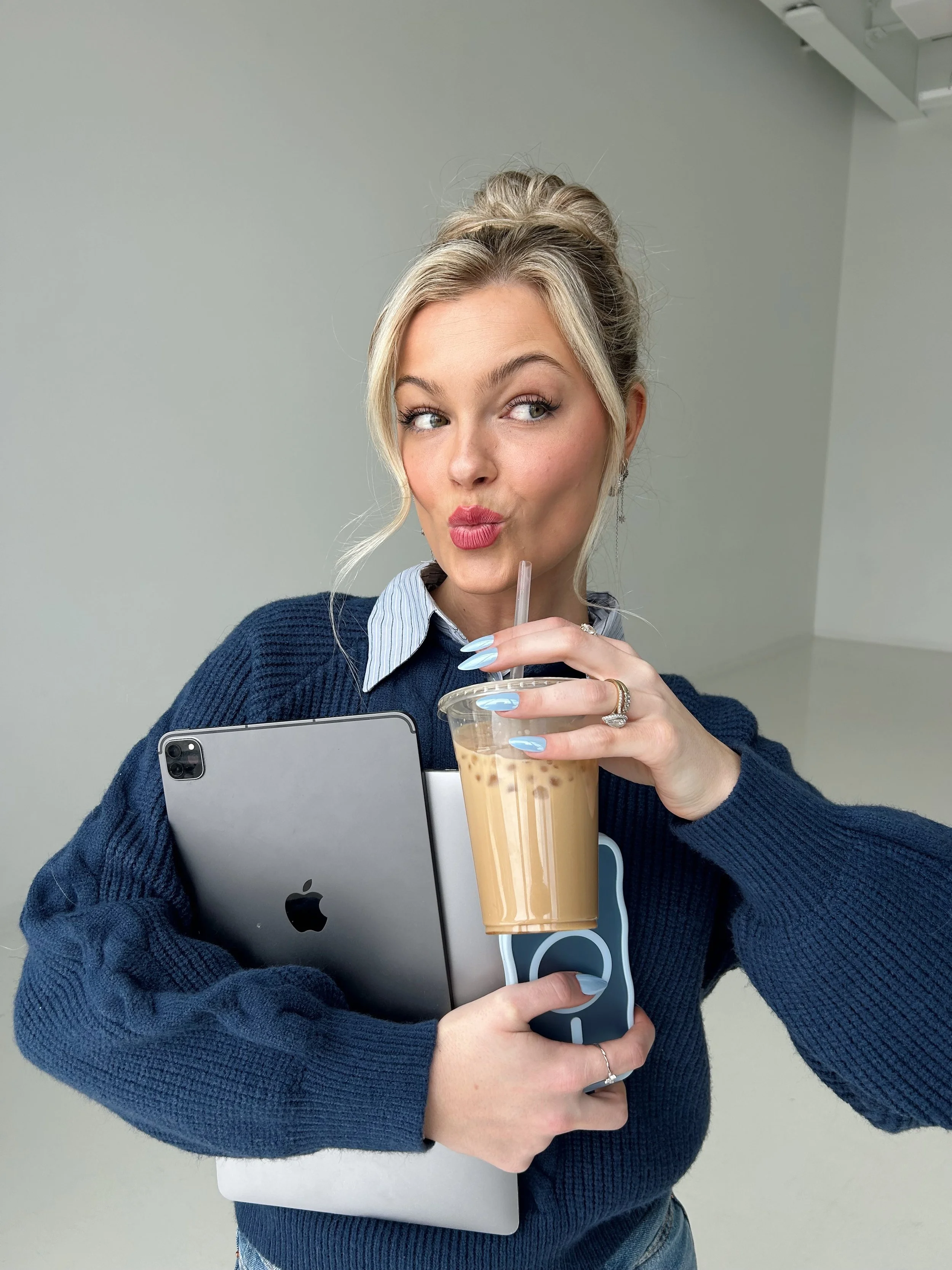 A young woman with blonde hair in a bun, wearing a blue sweater over a striped collared shirt, holding a tablet, ice coffee in a plastic cup with a straw, and a smartphone, while making a playful pouting face.
