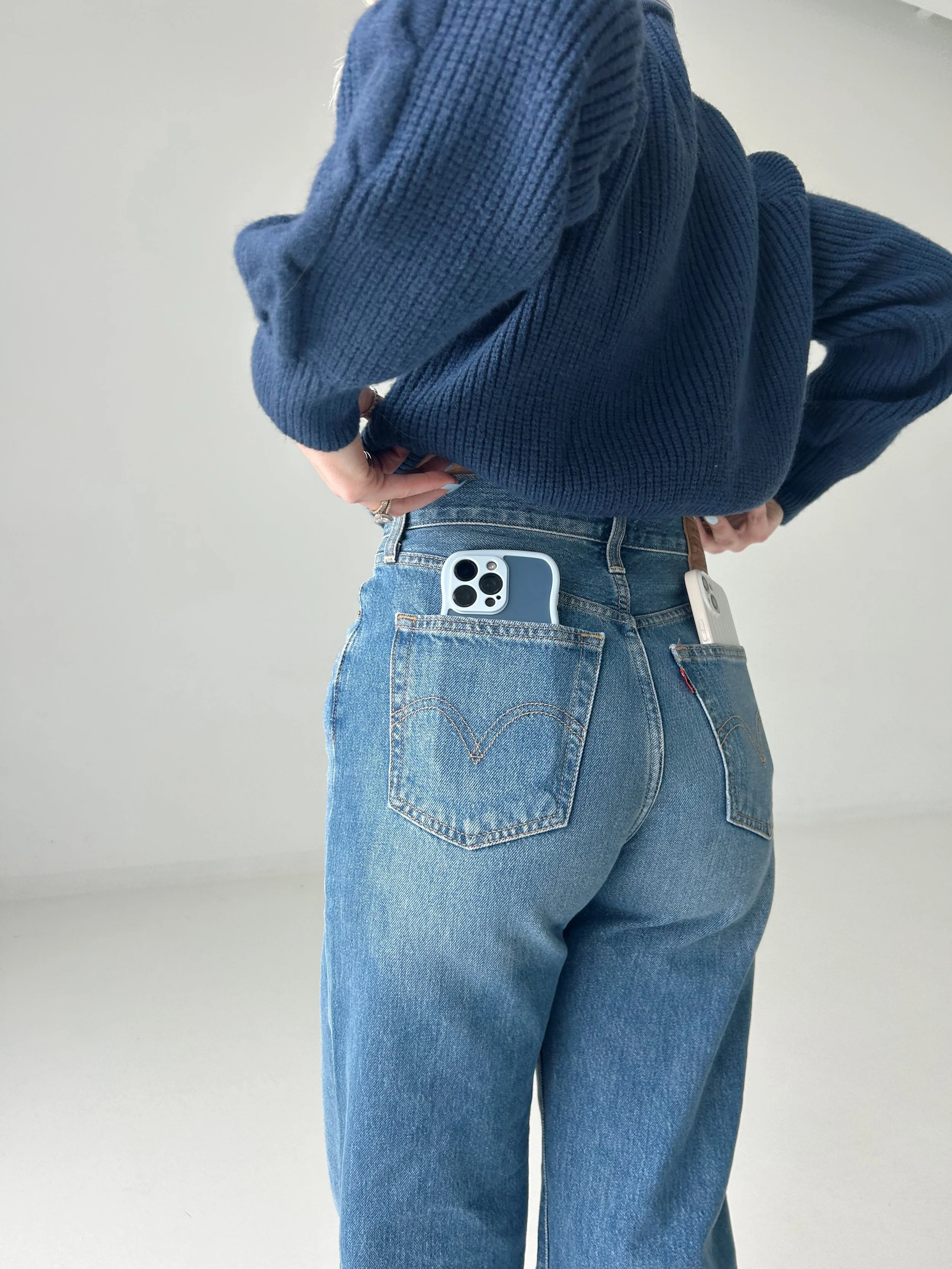 A person wearing blue jeans and a navy blue sweater with two smartphones in the back pockets.