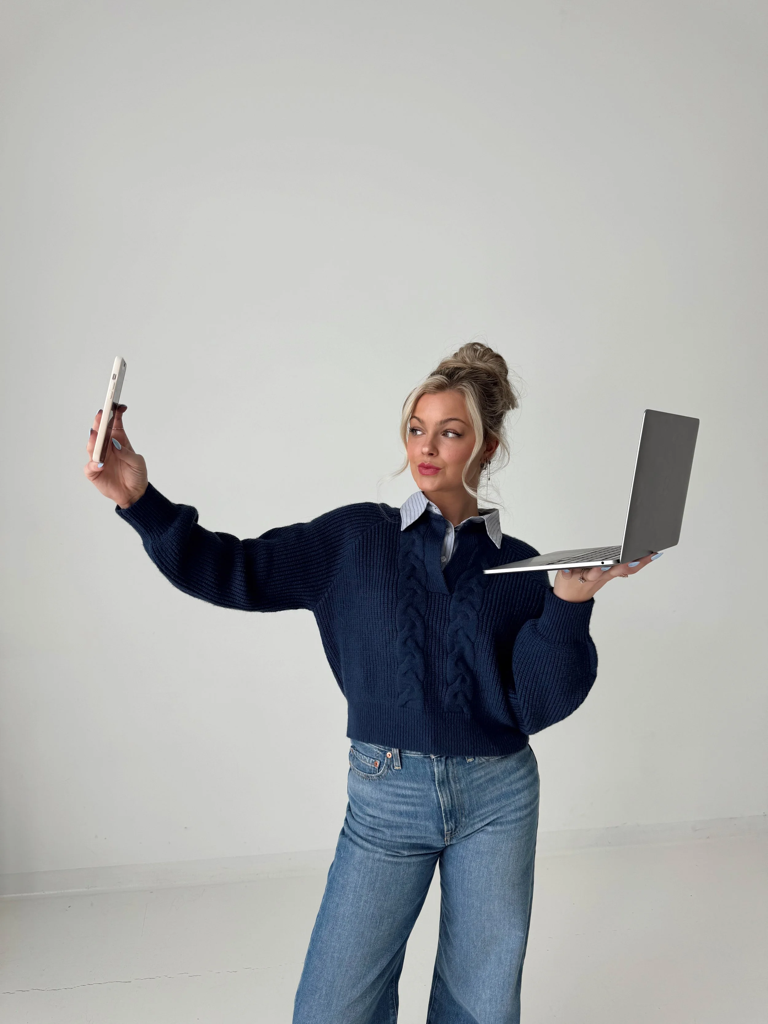 A young woman with blonde hair in a messy bun, wearing a navy blue sweater and jeans, taking a selfie with her smartphone in one hand while holding a laptop in the other.