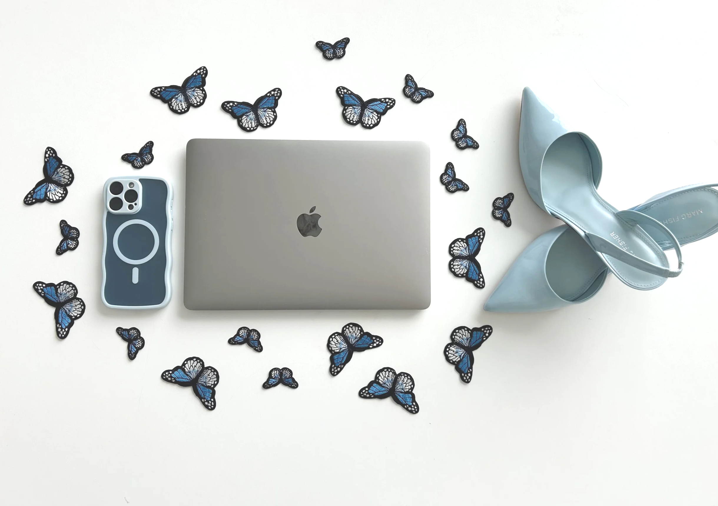 A flat lay photo of a closed silver MacBook, a blue and gray phone case, a pair of light blue high-heeled shoes, and black and blue butterfly cutouts on a white background.