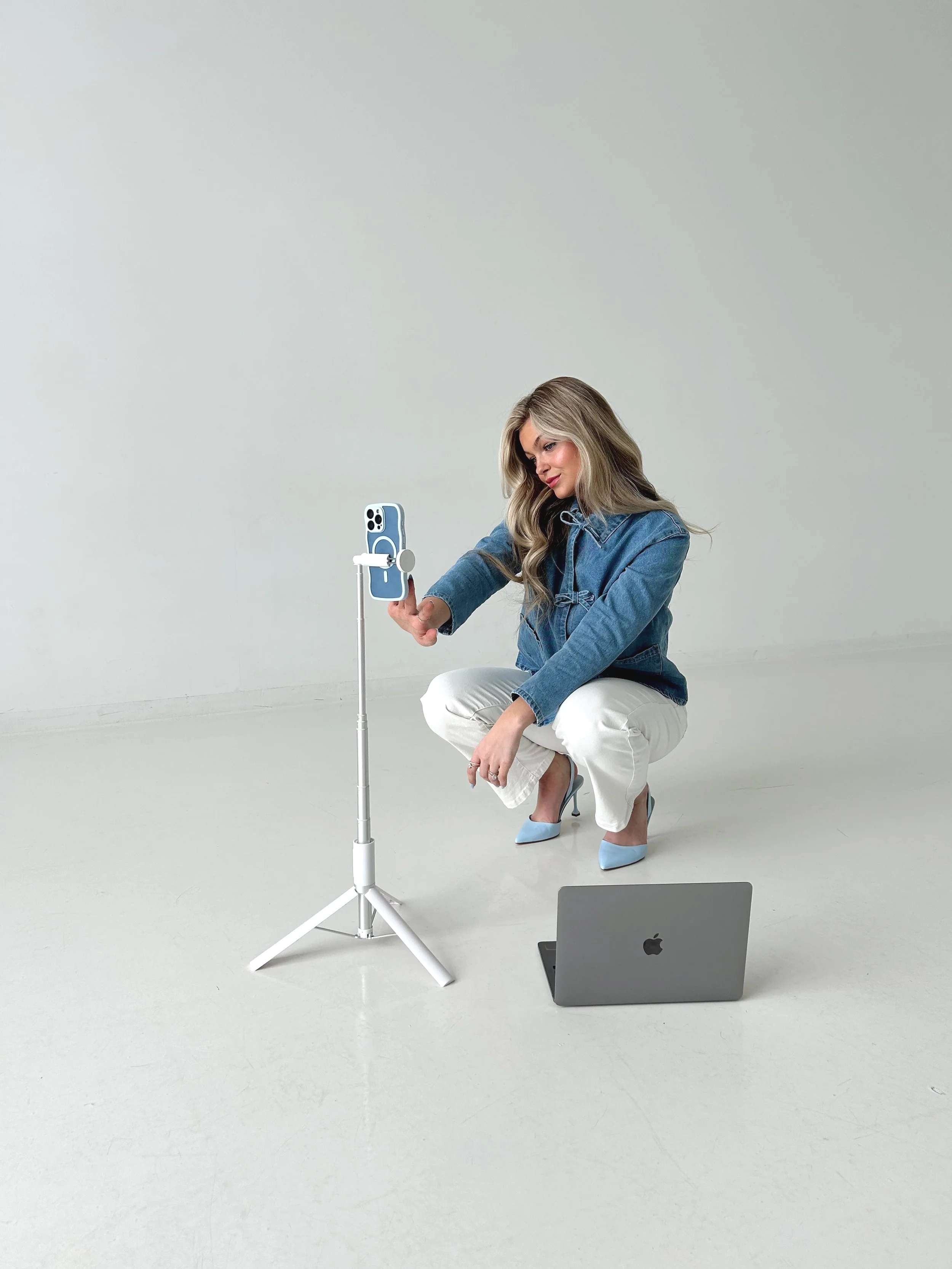 A woman crouching on the floor, taking a selfie with her smartphone on a tripod, with a laptop on the ground beside her.