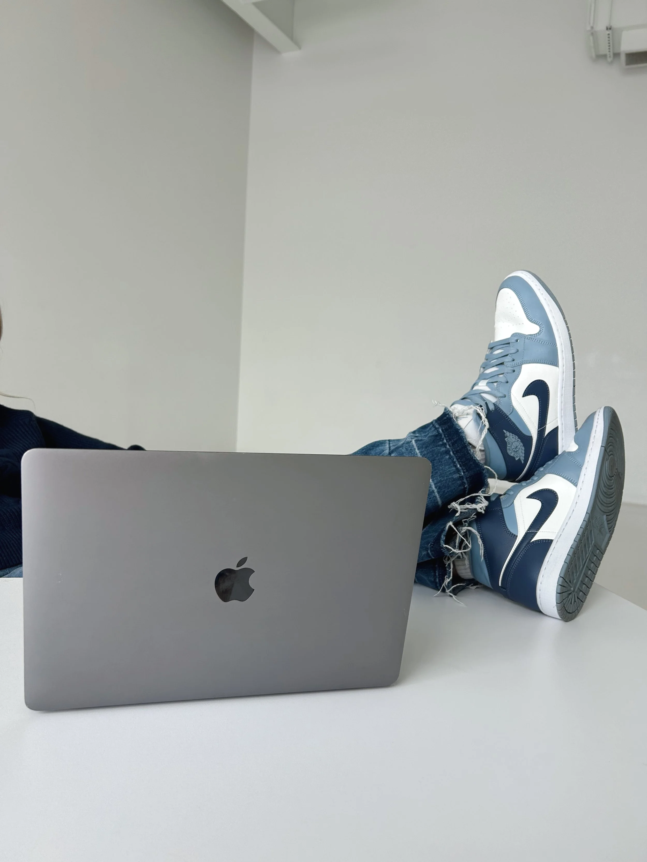 Person with legs extended on a white table, wearing blue and white sneakers and ripped jeans, with a closed silver MacBook on the table.