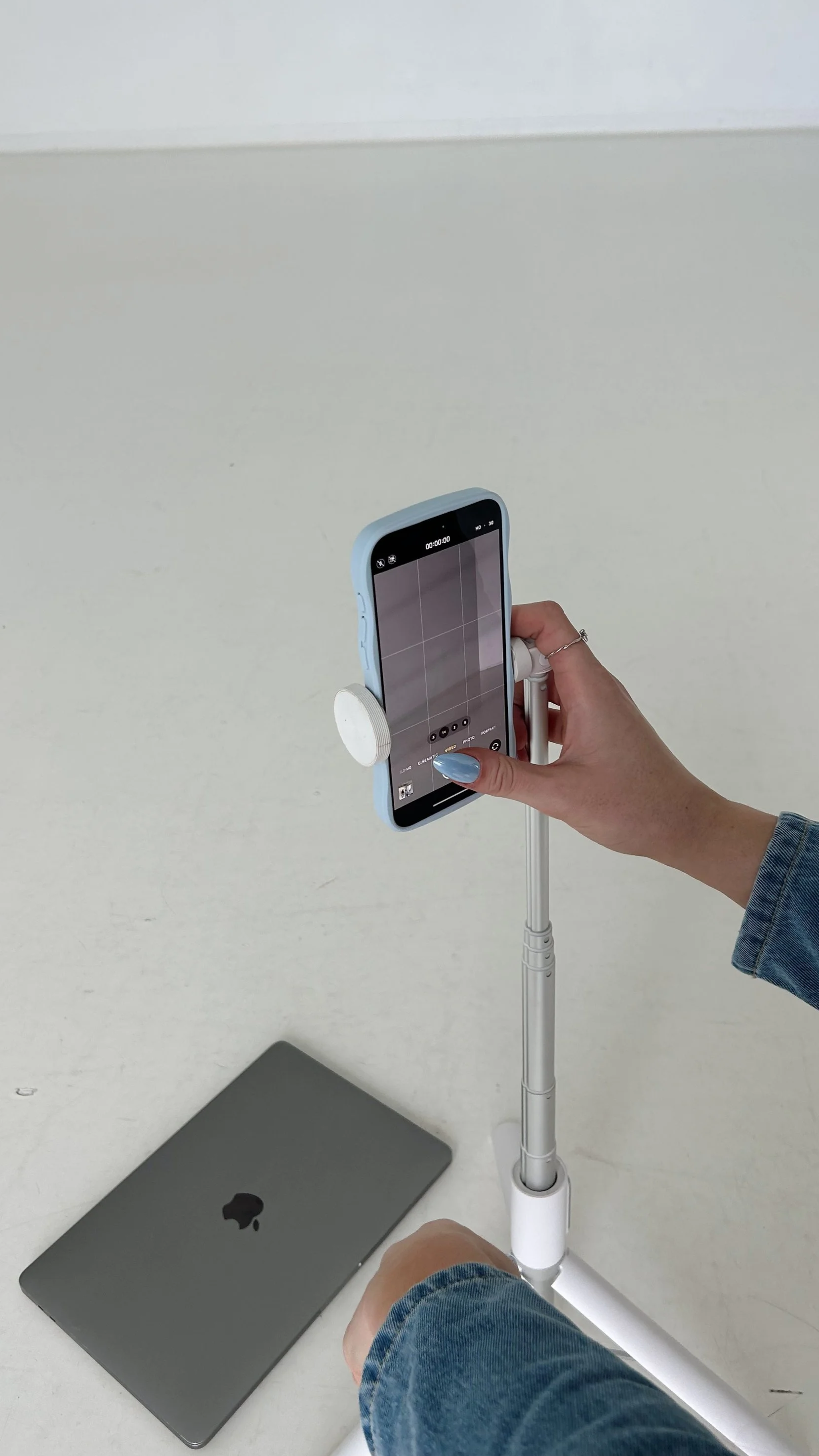 A person holding a smartphone mounted on a tripod, with a closed MacBook on a white surface nearby.