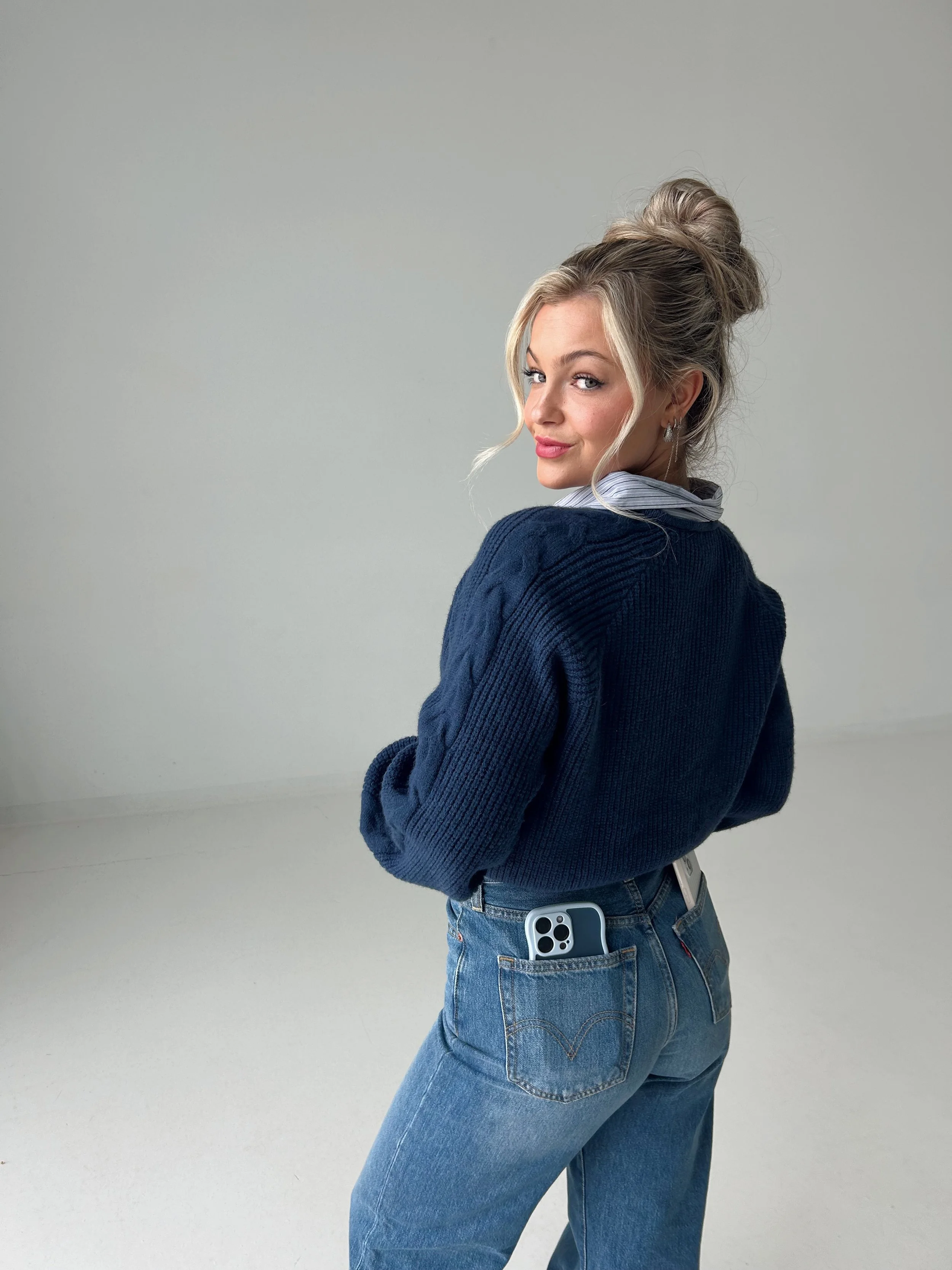 A young woman with blonde hair tied in a messy bun, wearing a navy blue sweater and jeans, standing with her back turned and looking over her shoulder, with a smartphone in her back pocket.