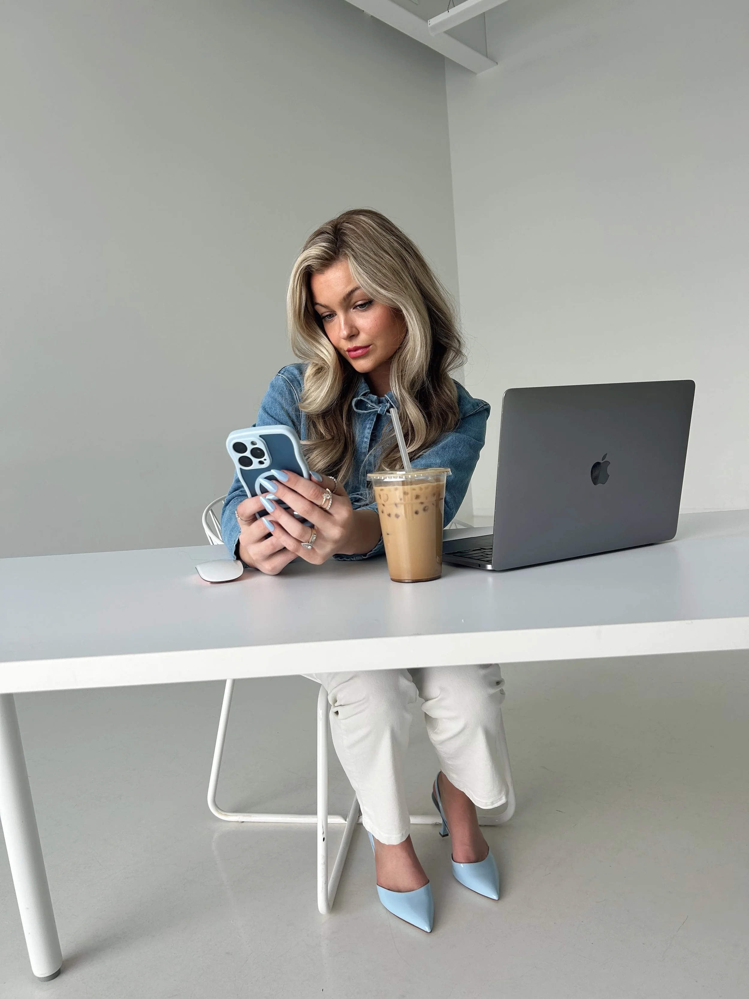 A woman sitting at a white table, looking at her smartphone, with a laptop and a glass of iced coffee in front of her. The setting is minimalistic with white walls and light-colored flooring.