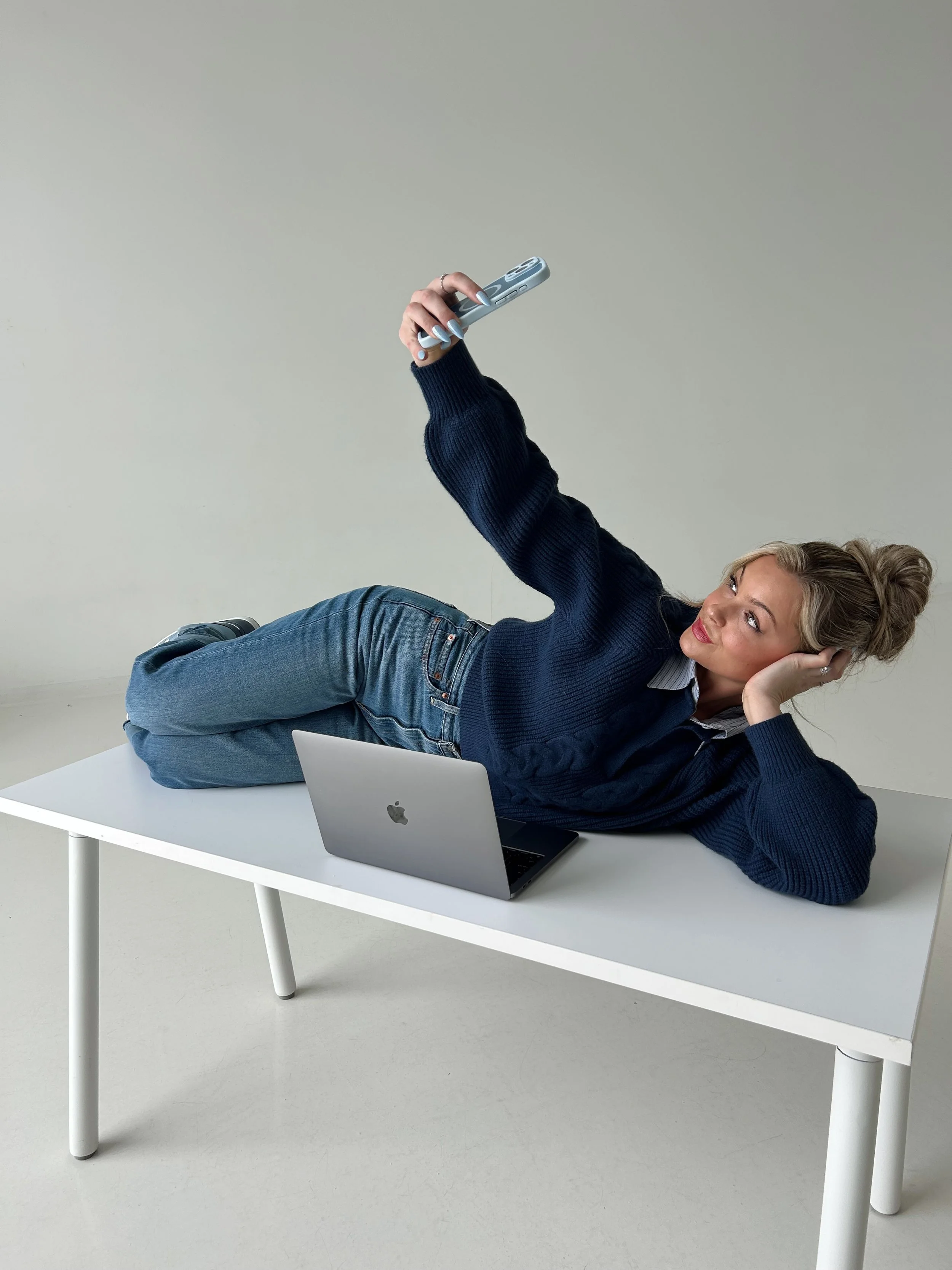 Woman lying on a white table with a laptop, taking a selfie with her smartphone, wearing a navy blue sweater and jeans, against a plain background.