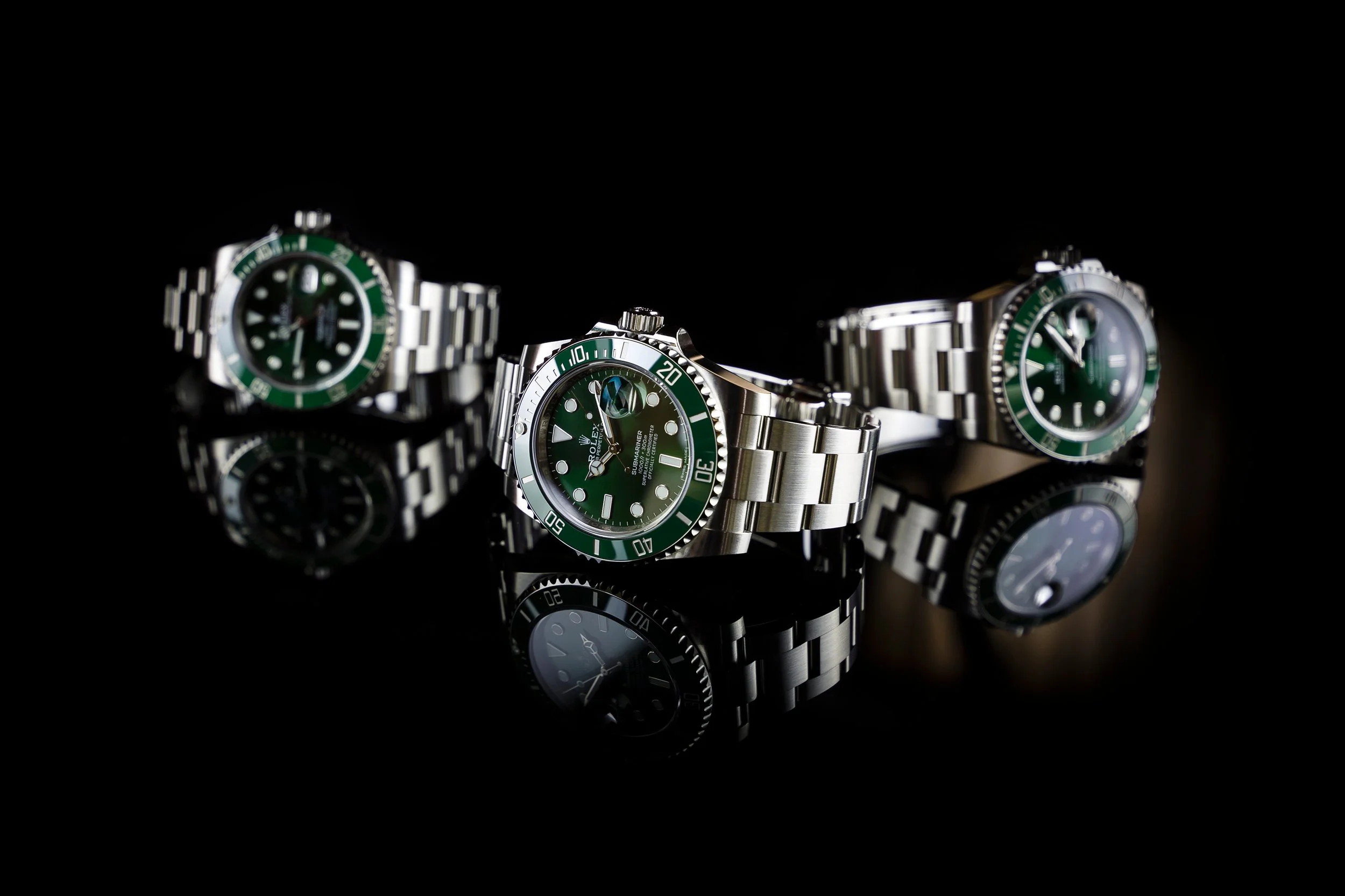 Three luxury Rolex watches with green faces and metal bracelets, displayed on a reflective black surface.