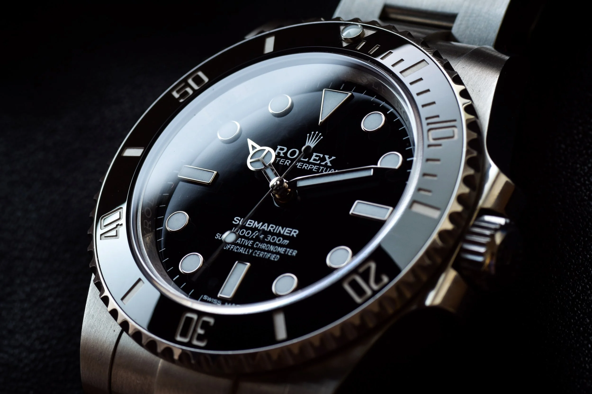 Close-up of a black and silver Rolex Submariner watch with a black dial, luminous markers, and a rotating bezel, on a black background.