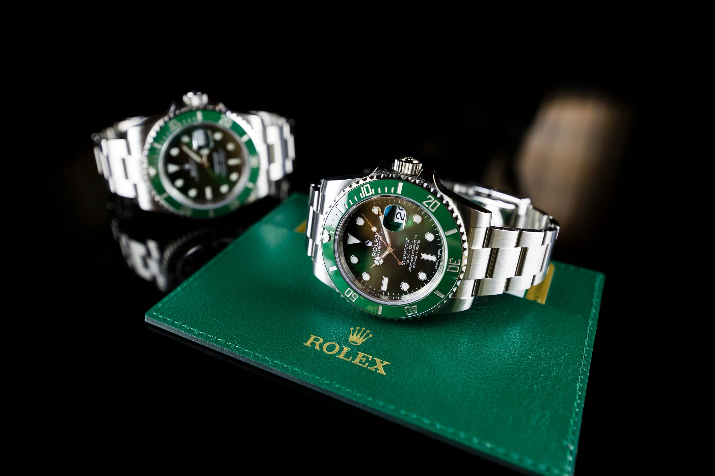 Two Rolex Submariner watches with green bezels, one placed on a green Rolex box and the other behind it, set against a black background.