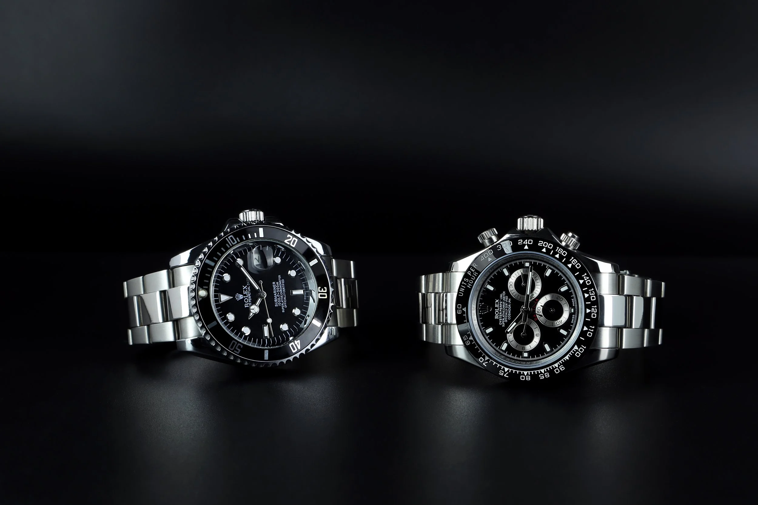 Two Rolex watches with black dials and stainless steel bracelets on a black surface against a black background.