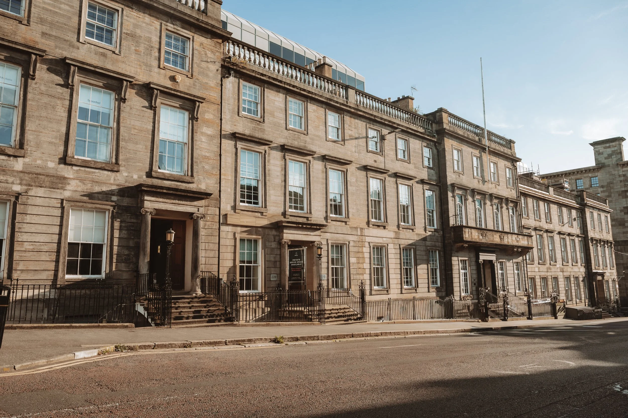 A historic multi-story building with stone exterior walls, numerous large windows, and steps leading up to entrances, located along a quiet street with a clear sky overhead.
