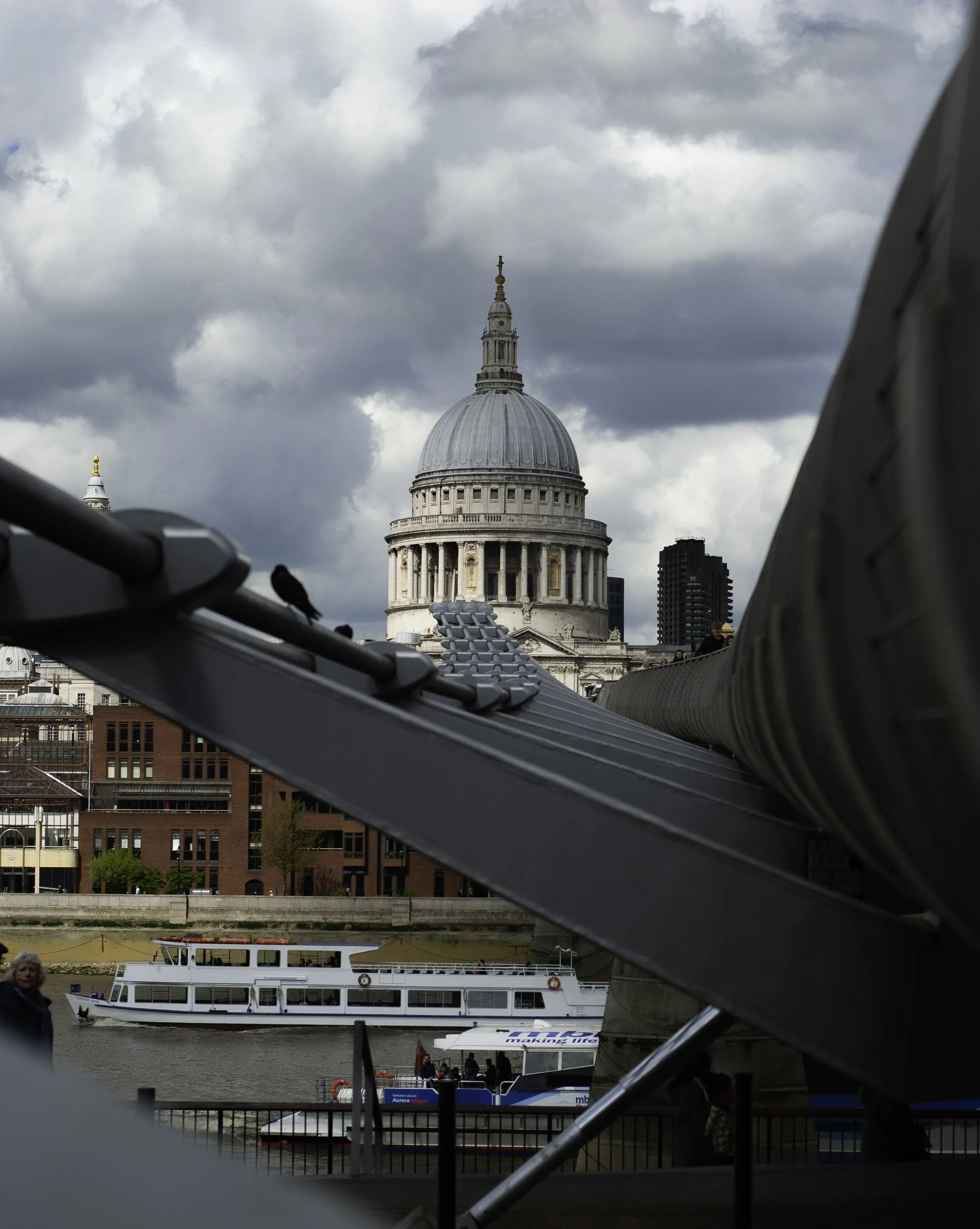 View of St. Paul's Cathedral in London, with boats on the river Thames, partly obscured by a bridge or structure, under a cloudy sky.