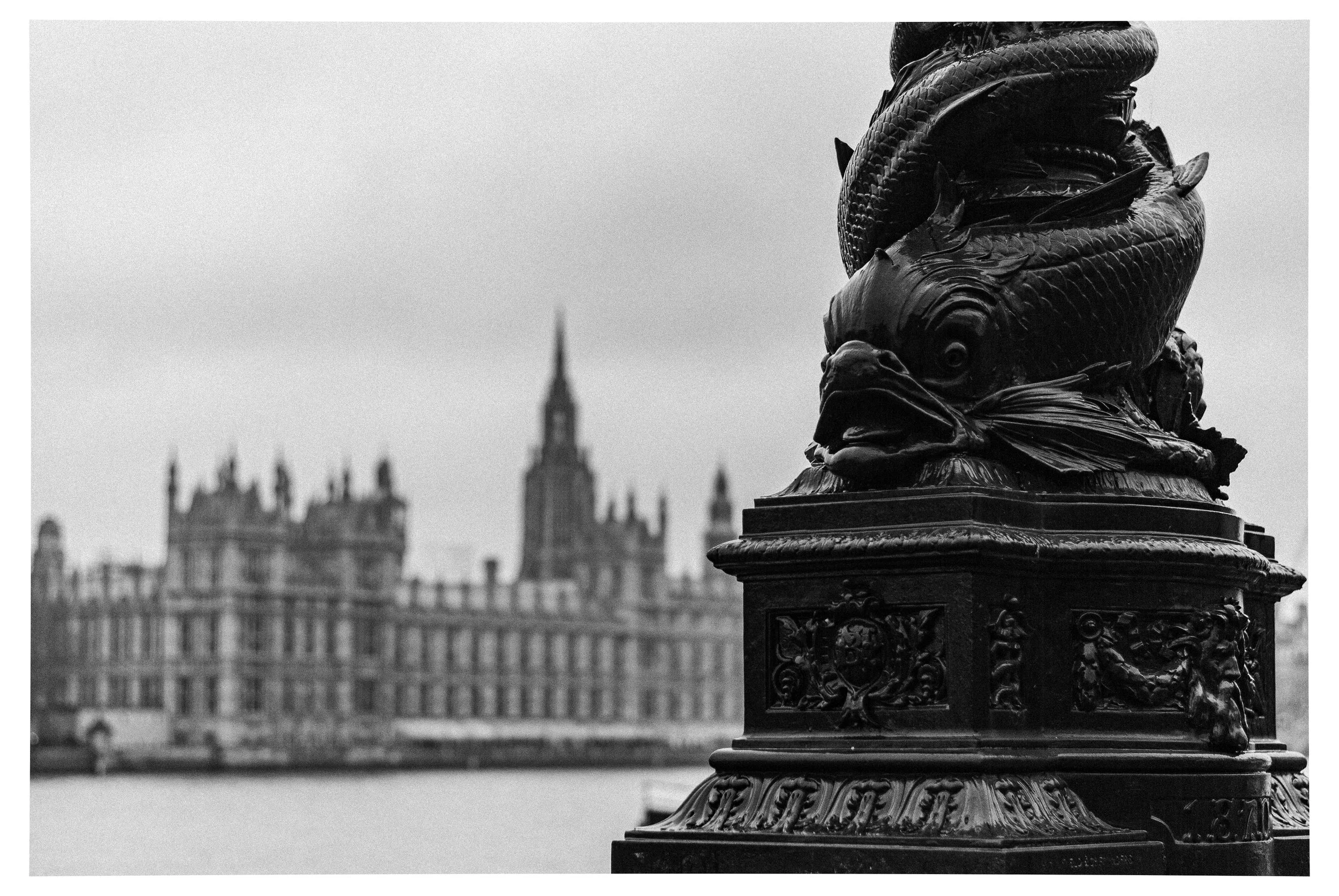 Black and white photo of a fish sculpture on a decorative pedestal with the Palace of Westminster in the background.