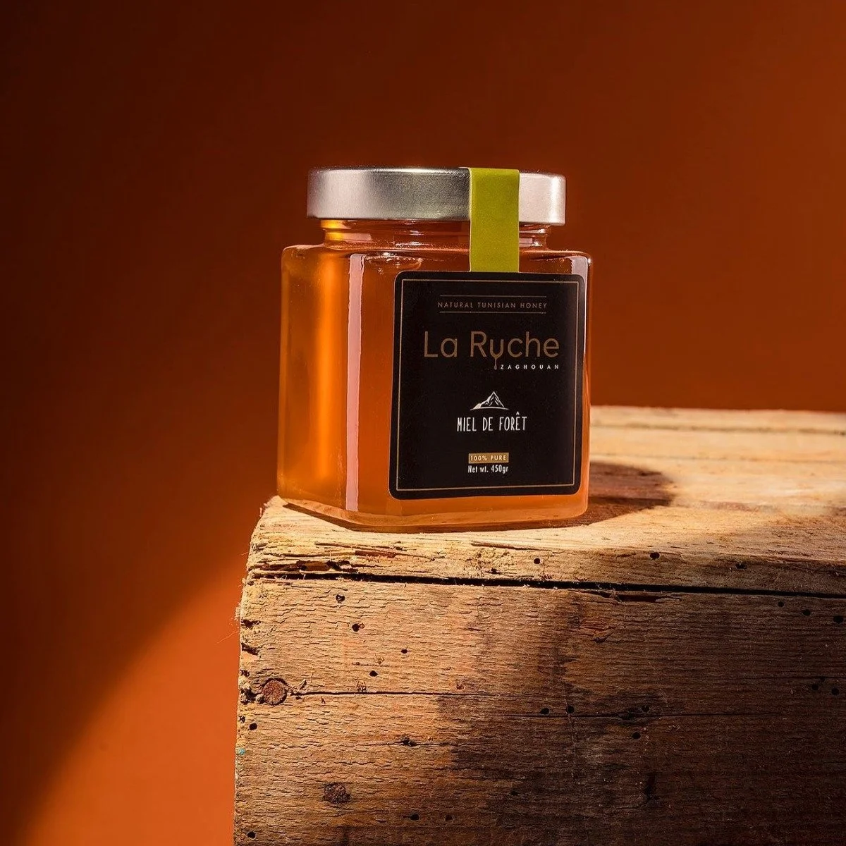 Jar of La Ruche honey labeled "Miel de Forêt" on a wooden surface with an orange background.
