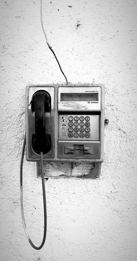 Black and white photo of an old wall-mounted payphone with a keypad, handset, and coin slot.