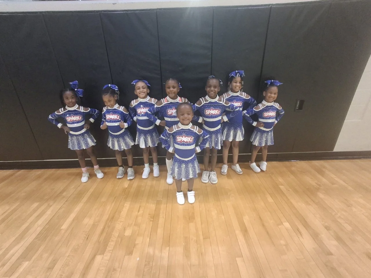 Group of eight young cheerleaders in blue and silver uniforms standing on a wooden gym floor against a black wall, smiling for the camera.
