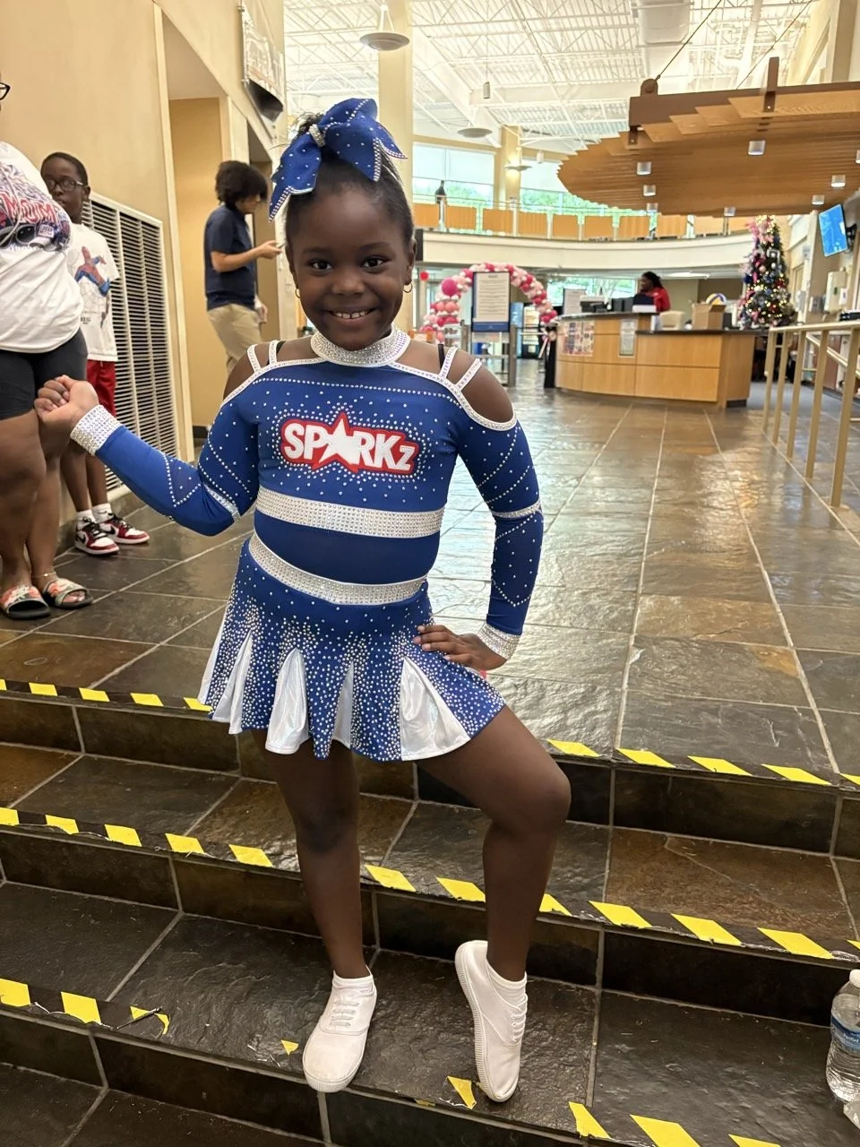 A young girl in a blue cheerleading outfit with white accents and crystals, standing on a tiled staircase inside a building, smiling and posing with one hand on her hip and the other raised in a fist. The outfit has a 'SPARKZ' logo on the front, and she has a matching blue bow in her hair, white shoes, and a choker. The background shows other children, a reception desk decorated for Christmas, and a Christmas tree.