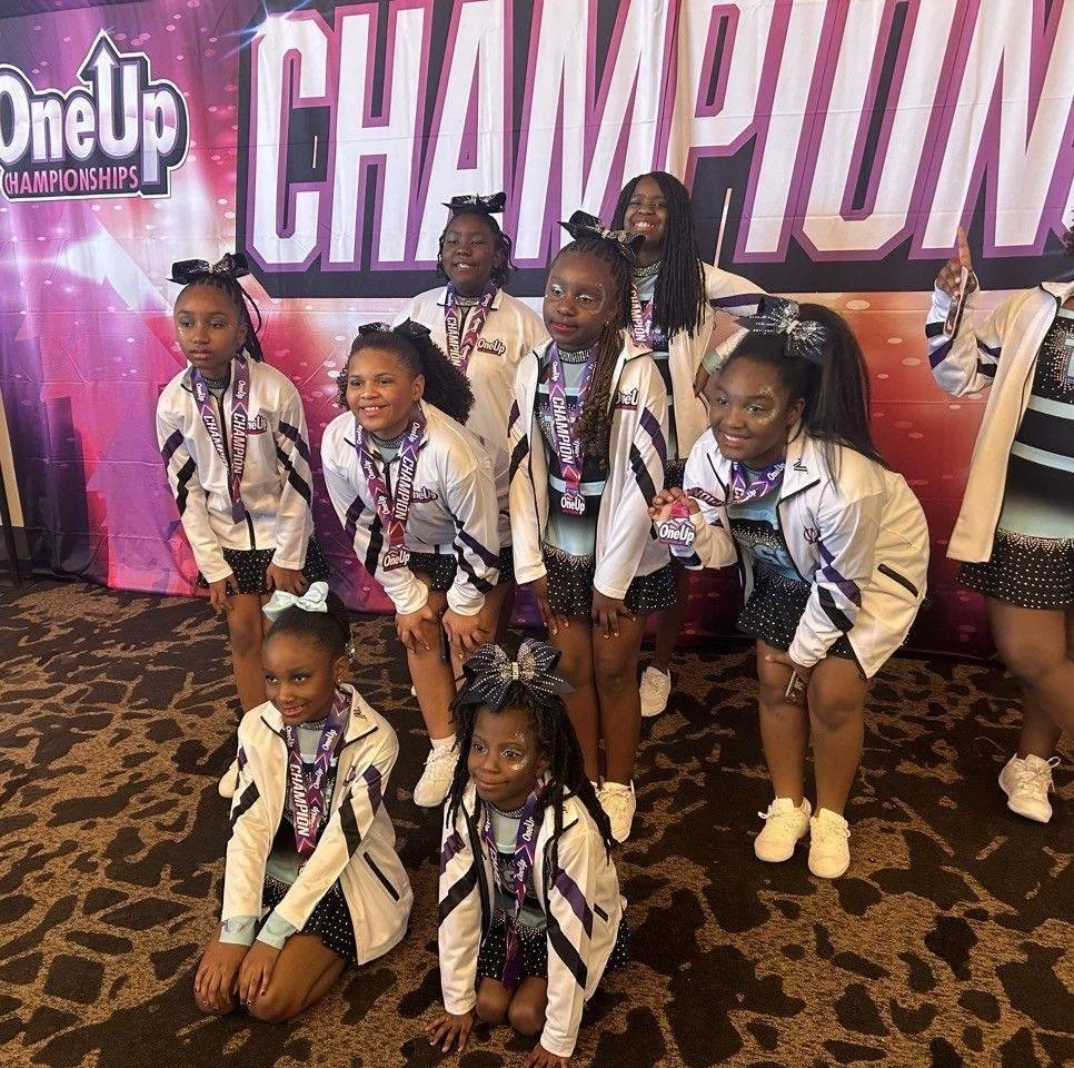 Group of young female cheerleaders dressed in white jackets and black skirts with polka dots celebrating their victory at the OneUp Cheer Championships, displaying medals and smiling, in front of a pink and purple backdrop with the words 'CHAMPION' in large letters.
