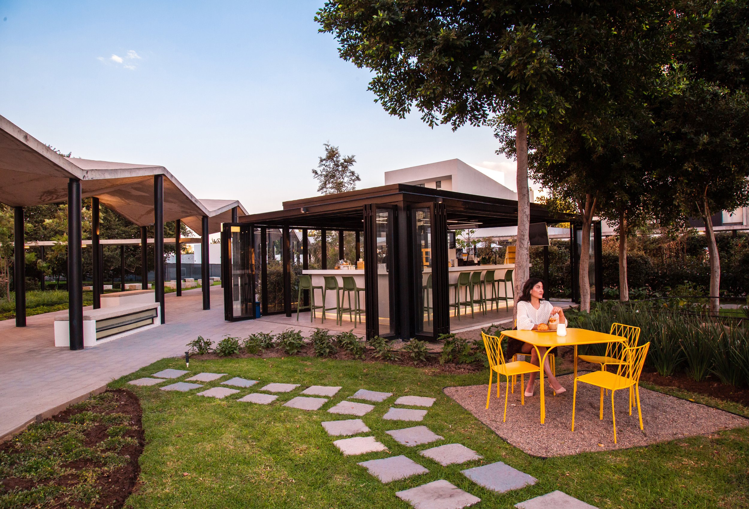 Una mujer sentada en una mesa amarilla en un jardín, con un quiosco de bar en el fondo, rodeado de árboles y césped.