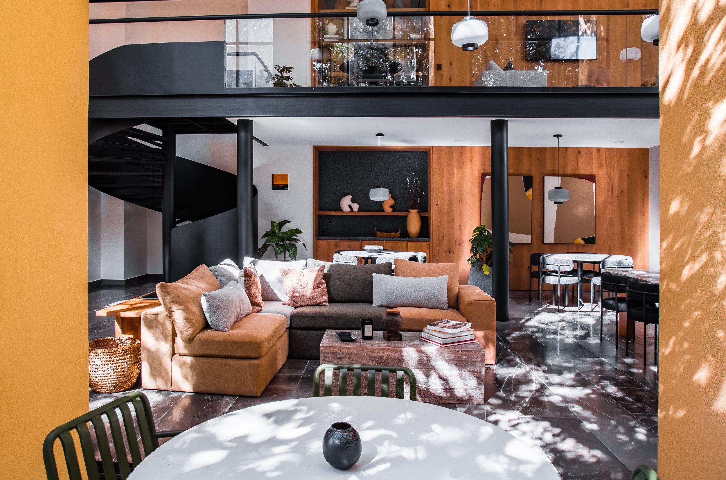 Interior de una sala de estar moderna con sofá en tonos crema y gris, mesa de centro de piedra, y paredes de madera,juega con iluminación natural y sombras de hojas. En el fondo, una zona con paredes blancas y negro, con plantas y decoración artística.