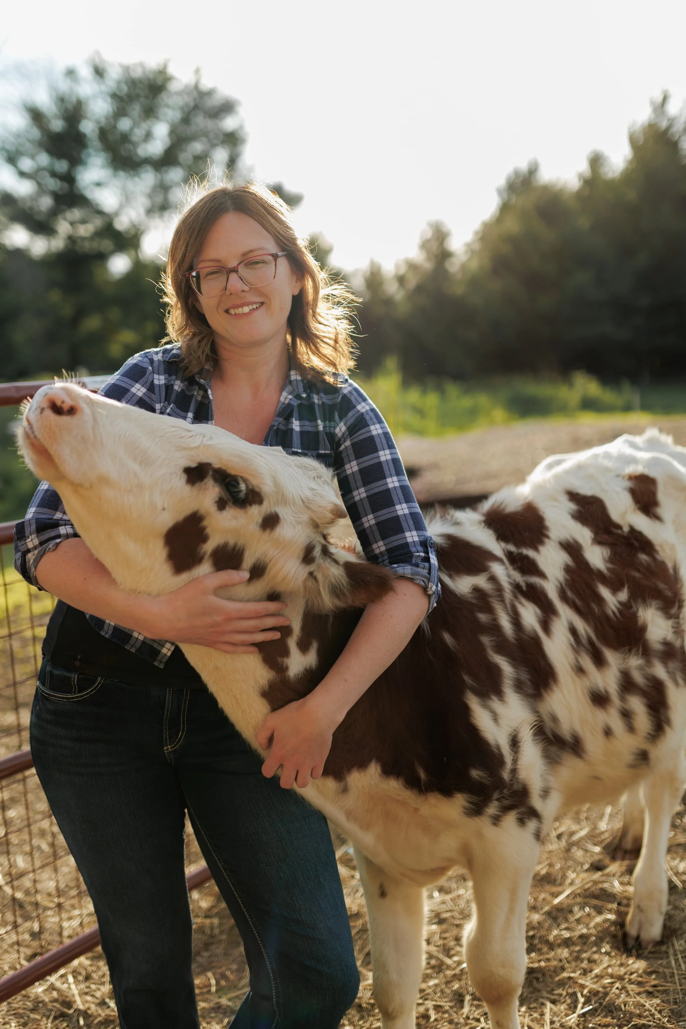 A woman with glasses and a plaid shirt smiling while holding a young calf with white and brown fur in a farm setting, with trees and a fence in the background.
