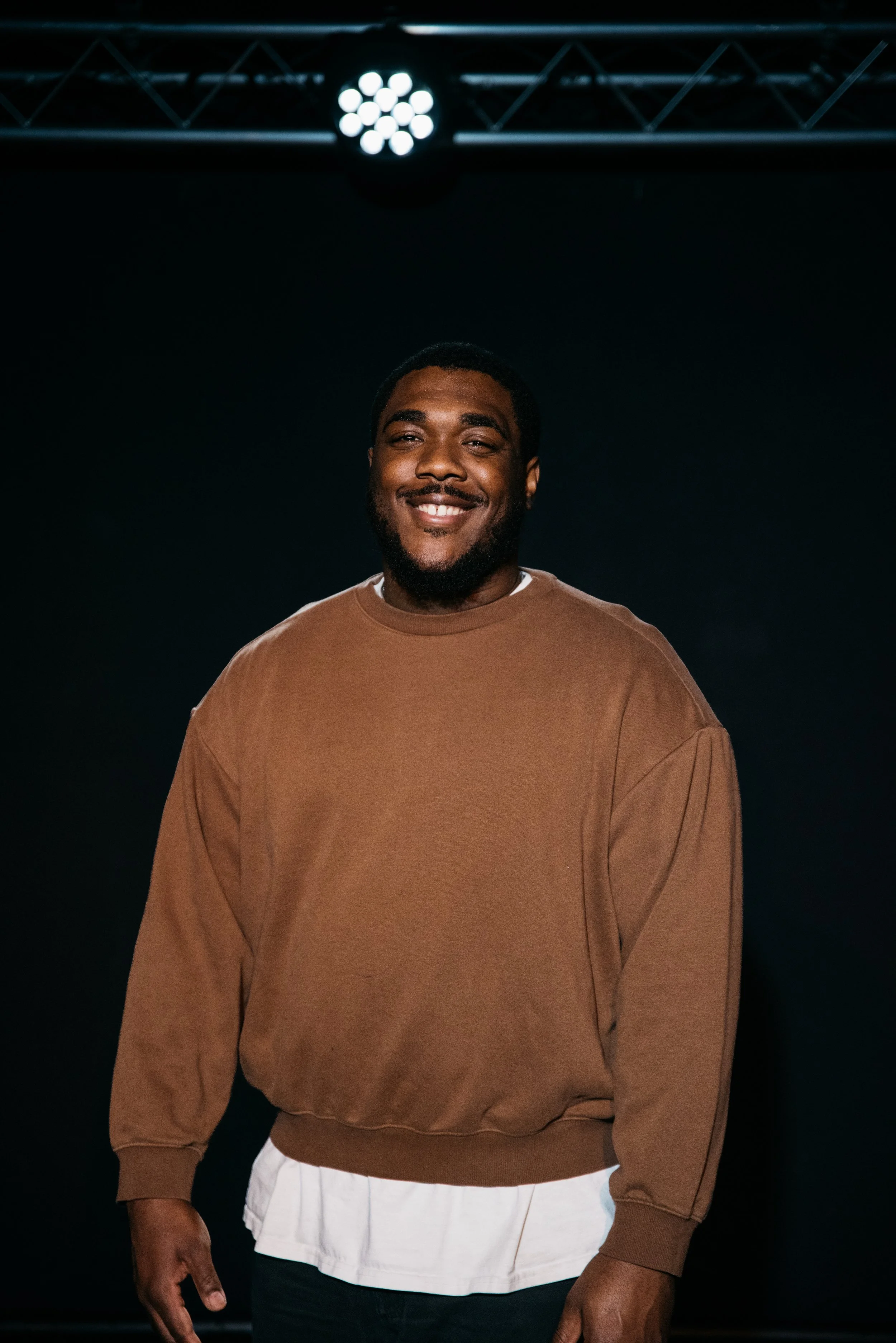 A smiling man with a beard and short hair, wearing a brown sweatshirt layered over a white shirt, standing in a dark space with stage lighting above.