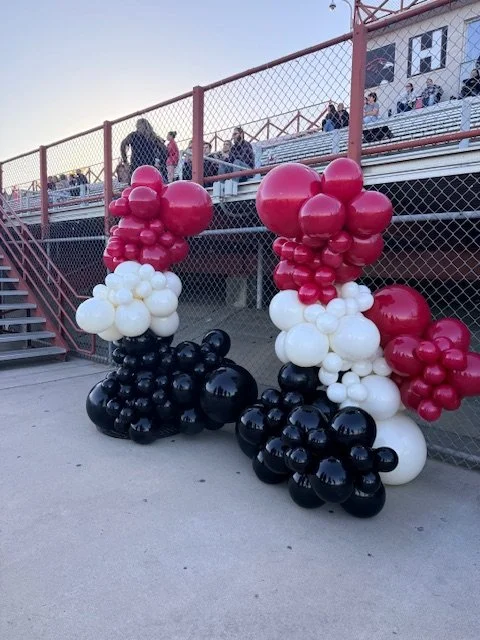 Balloon sculpture of a dog made with red, white, and black balloons, positioned on the ground near a chain-link fence and bleachers with spectators.
