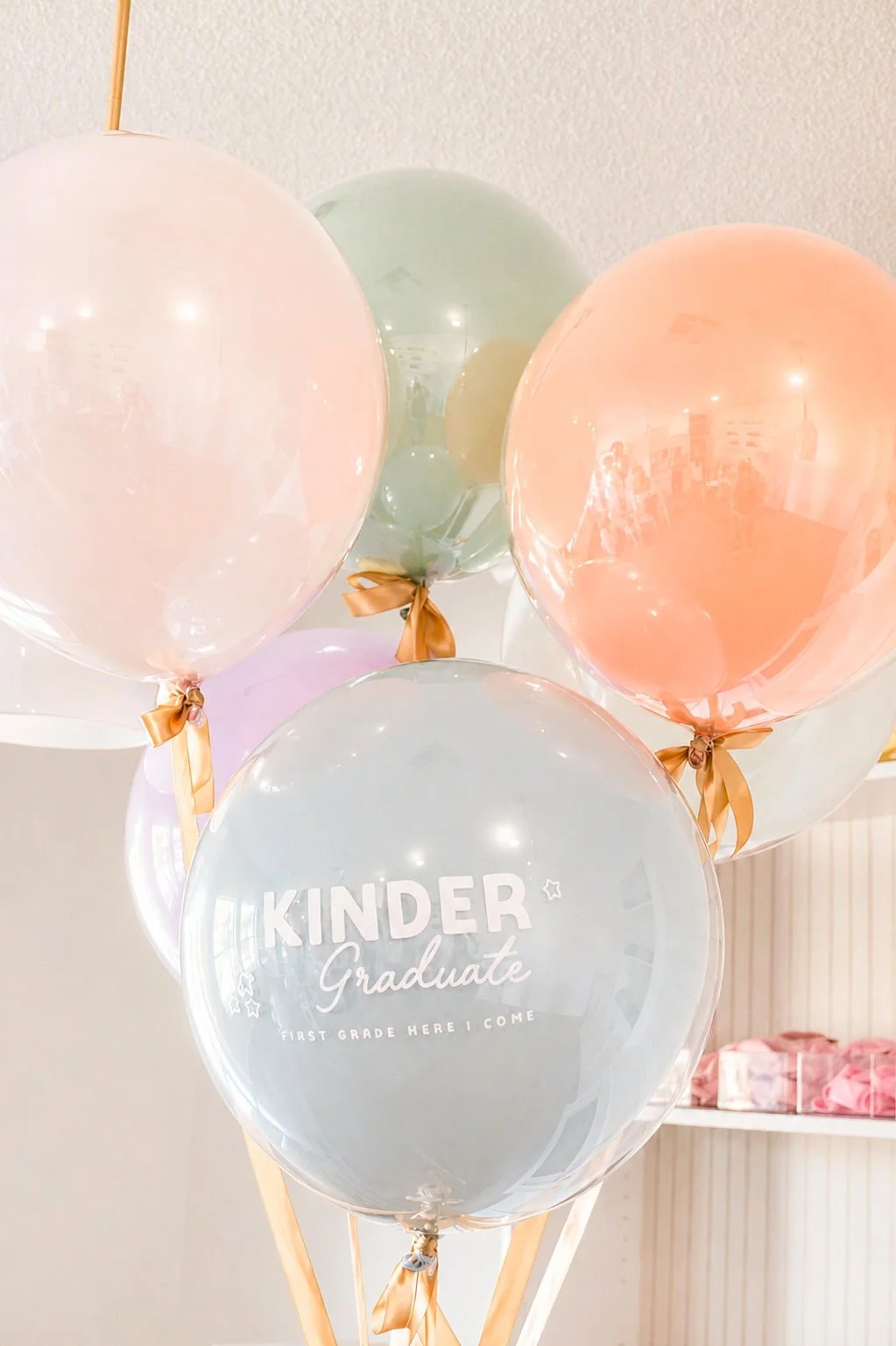 A cluster of pastel-colored balloons, including pink, green, purple, white, and peach, with gold ribbons tied at the knots. One balloon has the text 'KINDER Graduate First Grade Here I Come' printed on it, indicating a graduation celebration for a yo