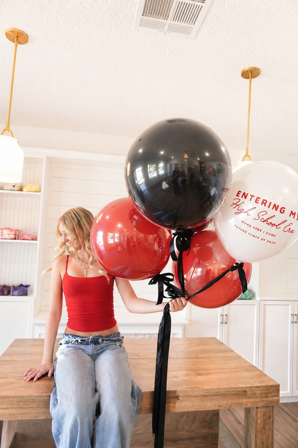 A young woman in a red tank top and baggy jeans sitting on a wooden table, holding a bunch of balloons, some of which have congratulatory messages about entering high school.