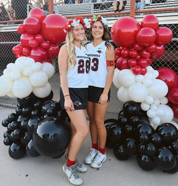 Two young women in sports jerseys and flower crowns standing in front of balloon decorations at a sporting event.