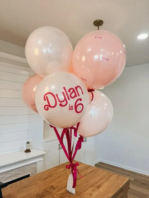 A bouquet of six helium balloons, some with pink and white colors, and one with the text 'Dylan is 6' in red lettering, tied with pink ribbons on a dining table.