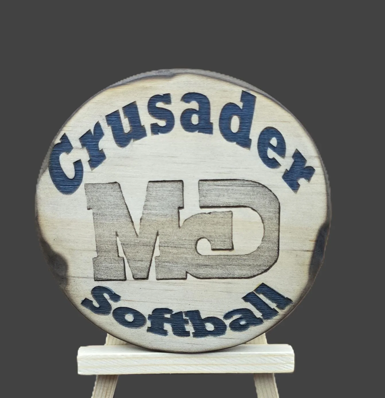 A wooden round sign with the words 'Genuine' at the top, 'softball' at the bottom, and a large 'GM' in the center. The sign is mounted on a small wooden easel.