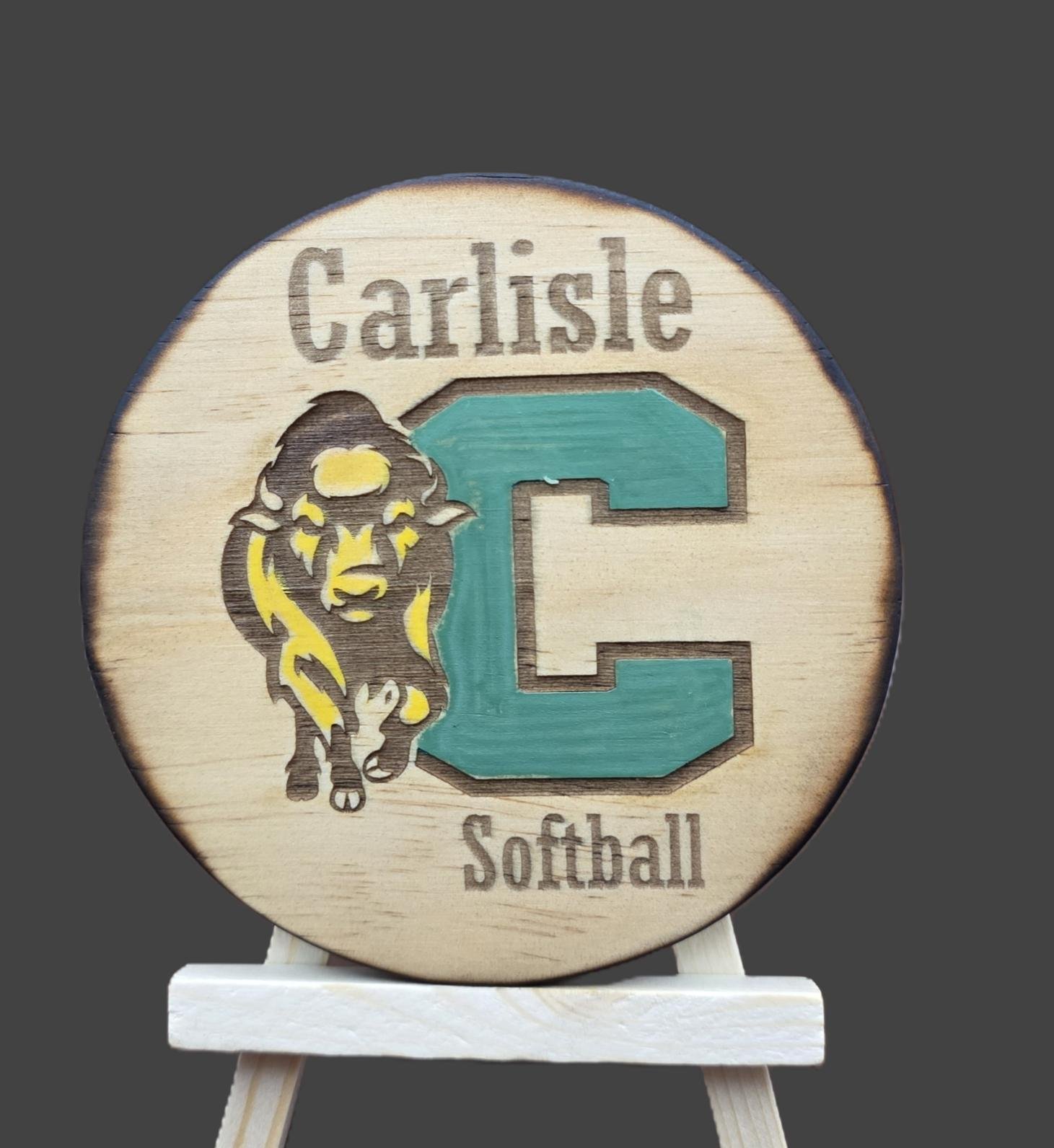 A round wooden sign featuring a caramel-colored background with dark edges, displaying a logo for Carlisle Softball. The logo includes a stylized yellow and black lion head with a teal letter 'C' next to it, and the words 'Carlisle' above and 'Softball' below.