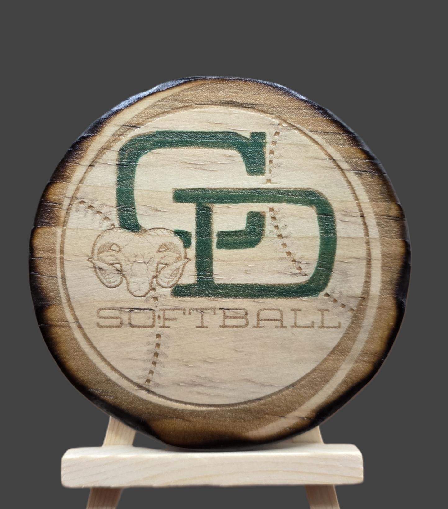 Wooden disc engraved with a logo that features a stylized letter combination and a ram's head, with the word 'SOFTBALL' below, displayed on a small easel against a dark background.