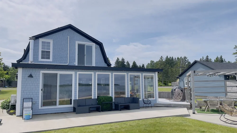 Waterfront side of a light blue cottage with dark trim and sunroom after an exterior repaint in Shediac